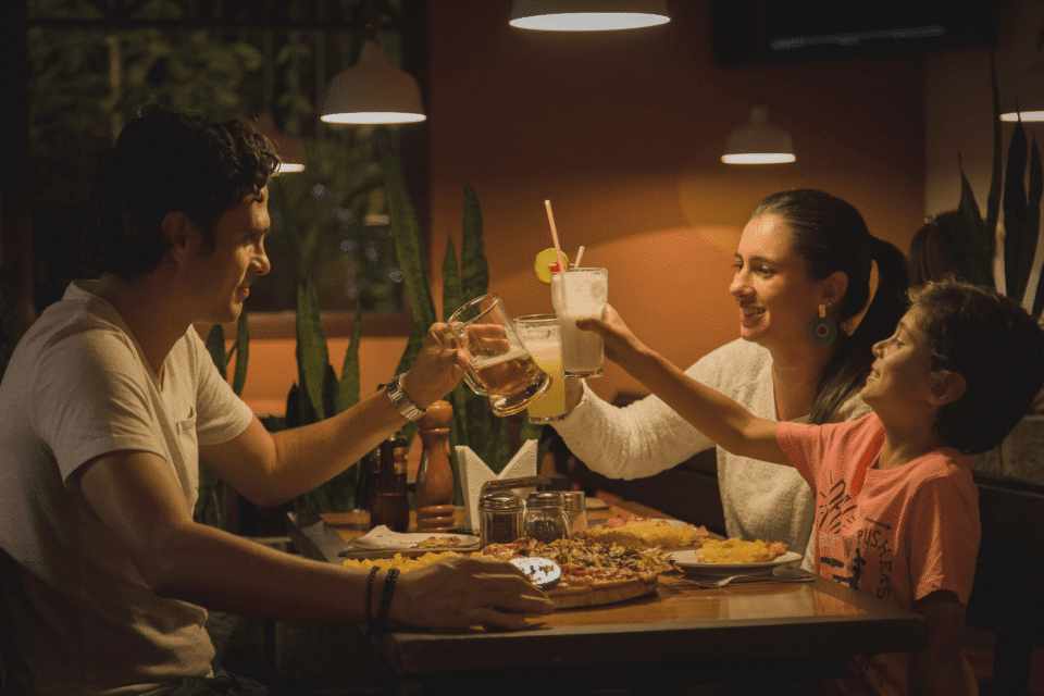 Een gezin geniet samen aan tafel met een Kieskeurige Eter van het eten, ze proosten hun glas boven de pizza die op tafel ligt. Je ziet een vader, moeder en kind.