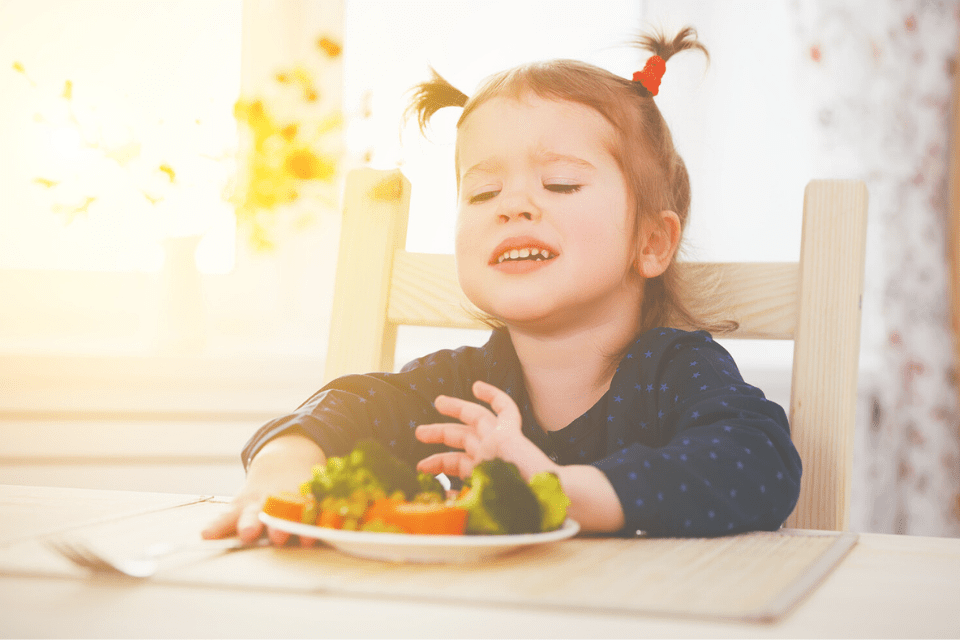 Kind dat bord weg schuift en ongelukkig kijkt omdat het geen groentes wil eten, kind zit aan tafel