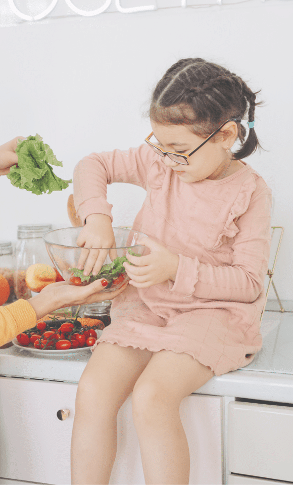 Een meisje zit op het aanrecht en houdt een glazen bak vast, waarin tomaatjes liggen. Met een hand pakt ze een tomaatje eruit. 