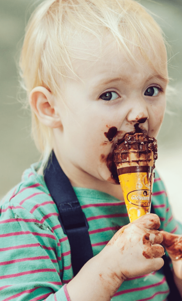 Een kindje met een ijsje vol in de mond, er zit een bruine kring om de mond van de chocoladesmaak. Het kind houdt het ijsje met 1 hand vast en kijkt in de camera.